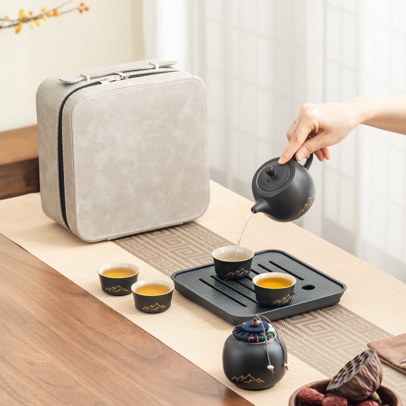 Black ceramic teapot pouring tea into one of four cups beside the square tray and cream carrying case