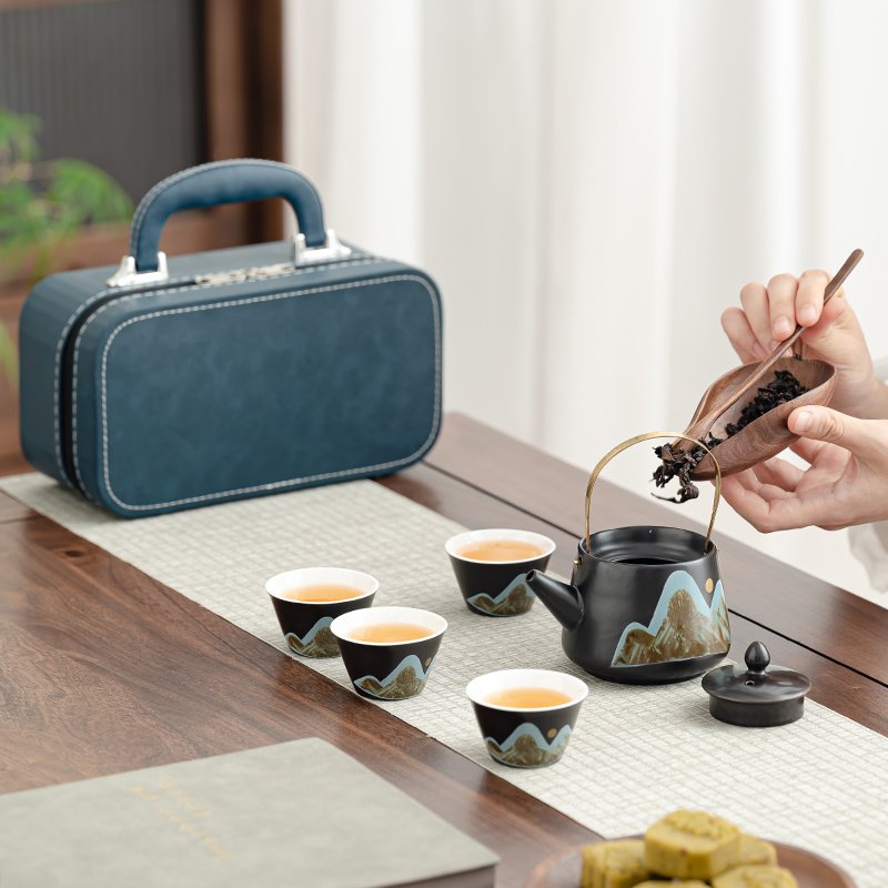 Loose tea being added to the open mountain-landscape teapot beside four cups and the blue carrying case