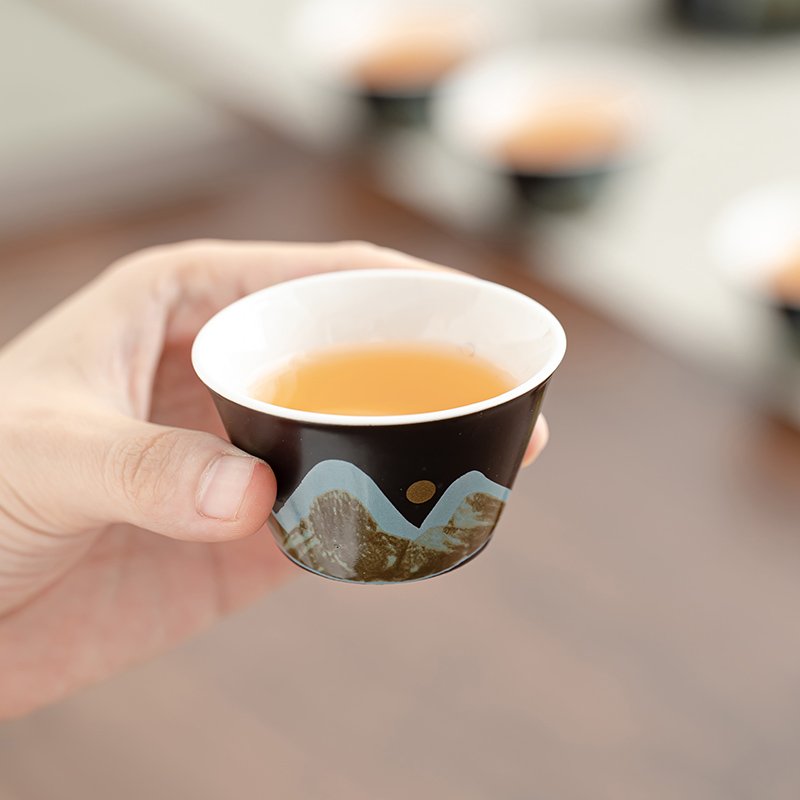 Small tea cup held in hand showing its light interior and blue and gold-tone mountain landscape
