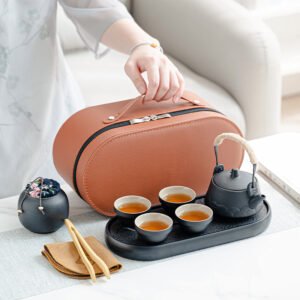 Black overhead-handle teapot and four cups on an oval tea tray beside a jar and brown carrying case